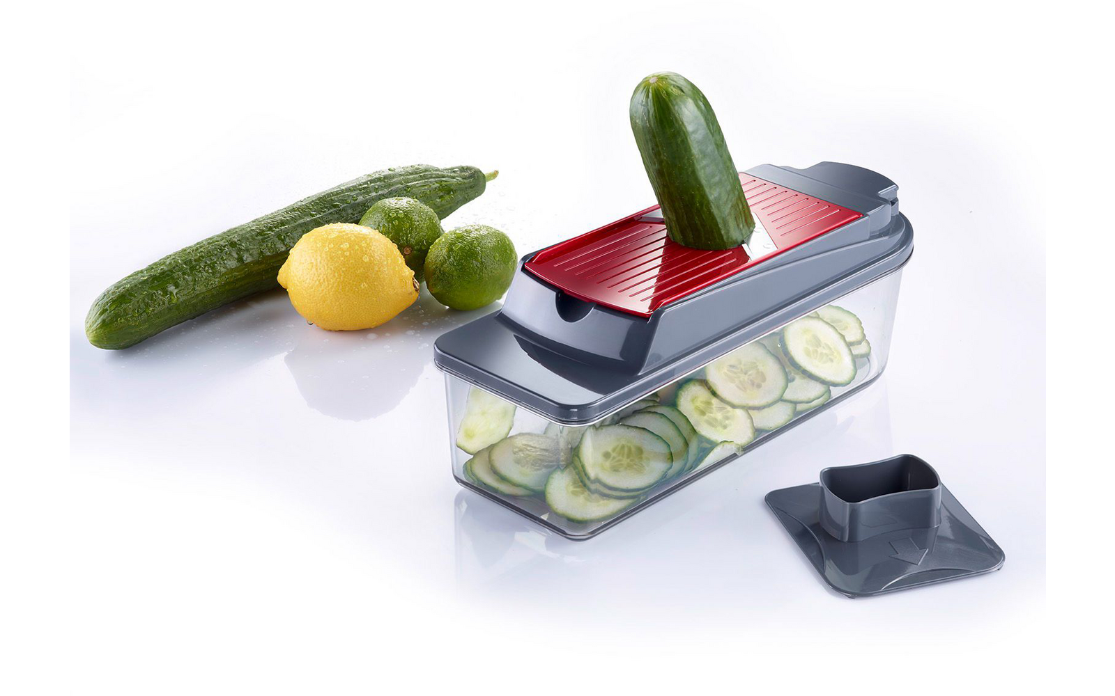Plastic vegetable slicer with red insert; a cucumber in the top opening; thin cucumber slices in the transparent container; two whole cucumbers, a lemon and a lime beside it; removable lid next to the container