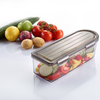 Transparent rectangular plastic container with sliced vegetables (zucchini, tomatoes, yellow and red bell pepper) in front; in the background a cucumber, green apple, lemon, red onion, two potatoes and a cluster of vine tomatoes