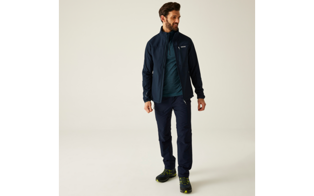 Mann steht, trägt dunkelblaue Outdoorjacke, blaugrünes T‑Shirt, dunkelblaue Hose und schwarze Wanderschuhe mit grünen Details; schaut nach rechts