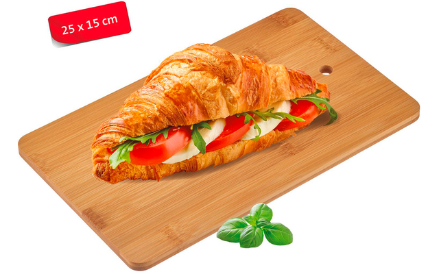 Croissant sandwich filled with tomato slices, white cheese slices and green leaves on a wooden cutting board; a few loose green leaves beside it; red label '25 x 15 cm' at top left