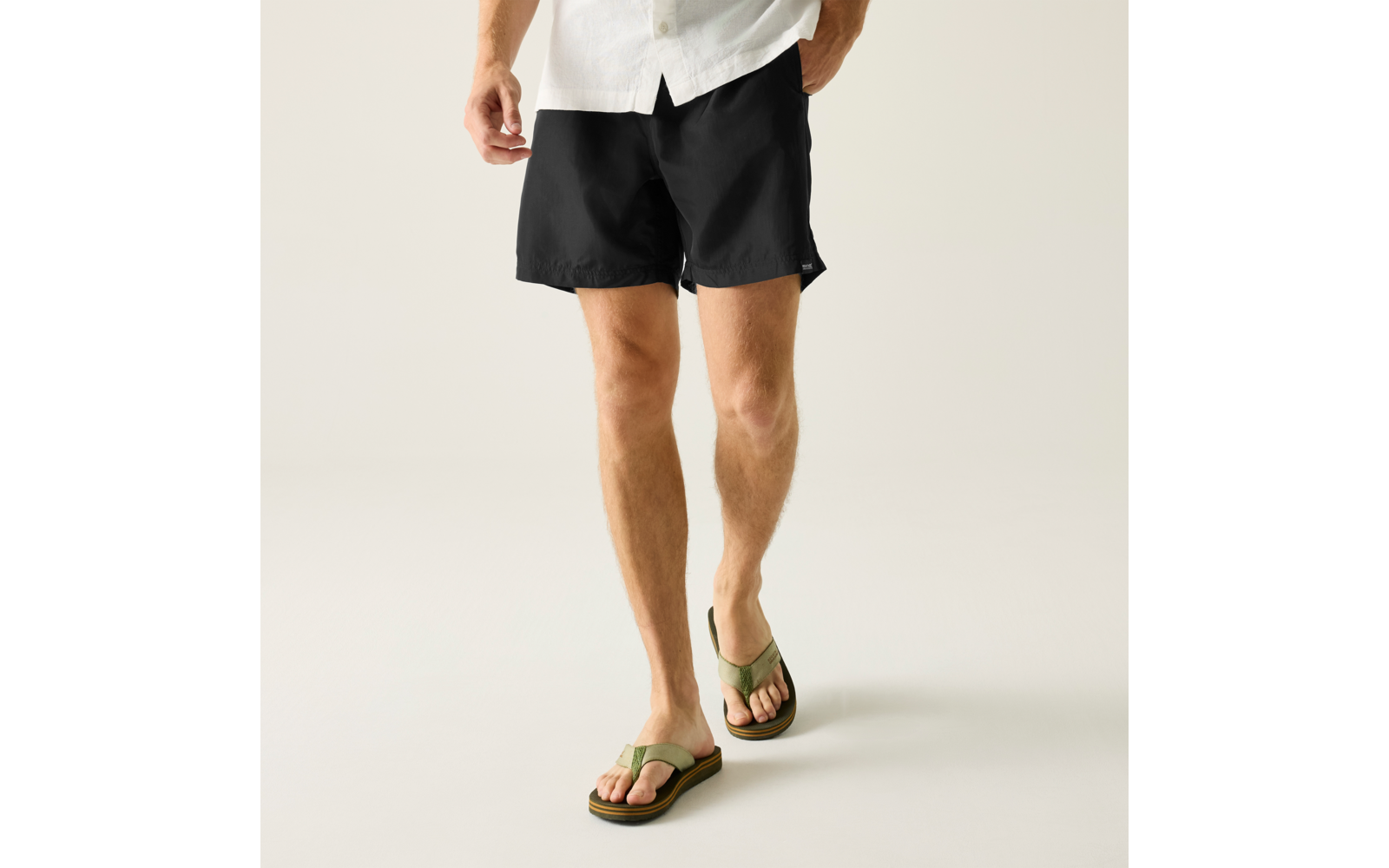 Hüftbereich bis Beine einer Person, weiße Hemdkante sichtbar, schwarze Shorts, ein Bein nach vorne gesetzt, grüne Zehensandalen, heller Hintergrund