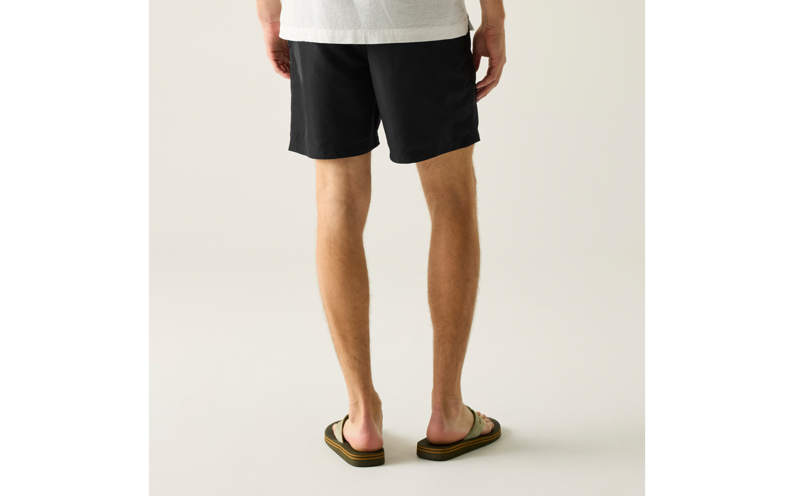 Rückenansicht der Beine und Hüfte einer Person, schwarze Shorts, weißer T-Shirt-Saum, grünliche Flip-Flops, heller neutraler Hintergrund