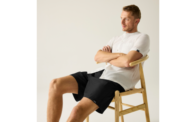 Mann sitzt auf einem Holzstuhl, Arme verschränkt, trägt weißes Hemd und schwarze Shorts, blickt nach rechts vor hellem Hintergrund