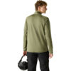 Personne vue de dos, porte une veste olive à manches longues et un pantalon noir, tient un casque noir dans la main droite, cheveux courts bouclés châtain clair, tête légèrement tournée à gauche