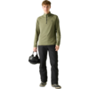 Personne debout portant un haut manches longues olive avec une courte fermeture éclair et un pantalon noir, tenant un casque noir dans la main droite sur un fond uni clair