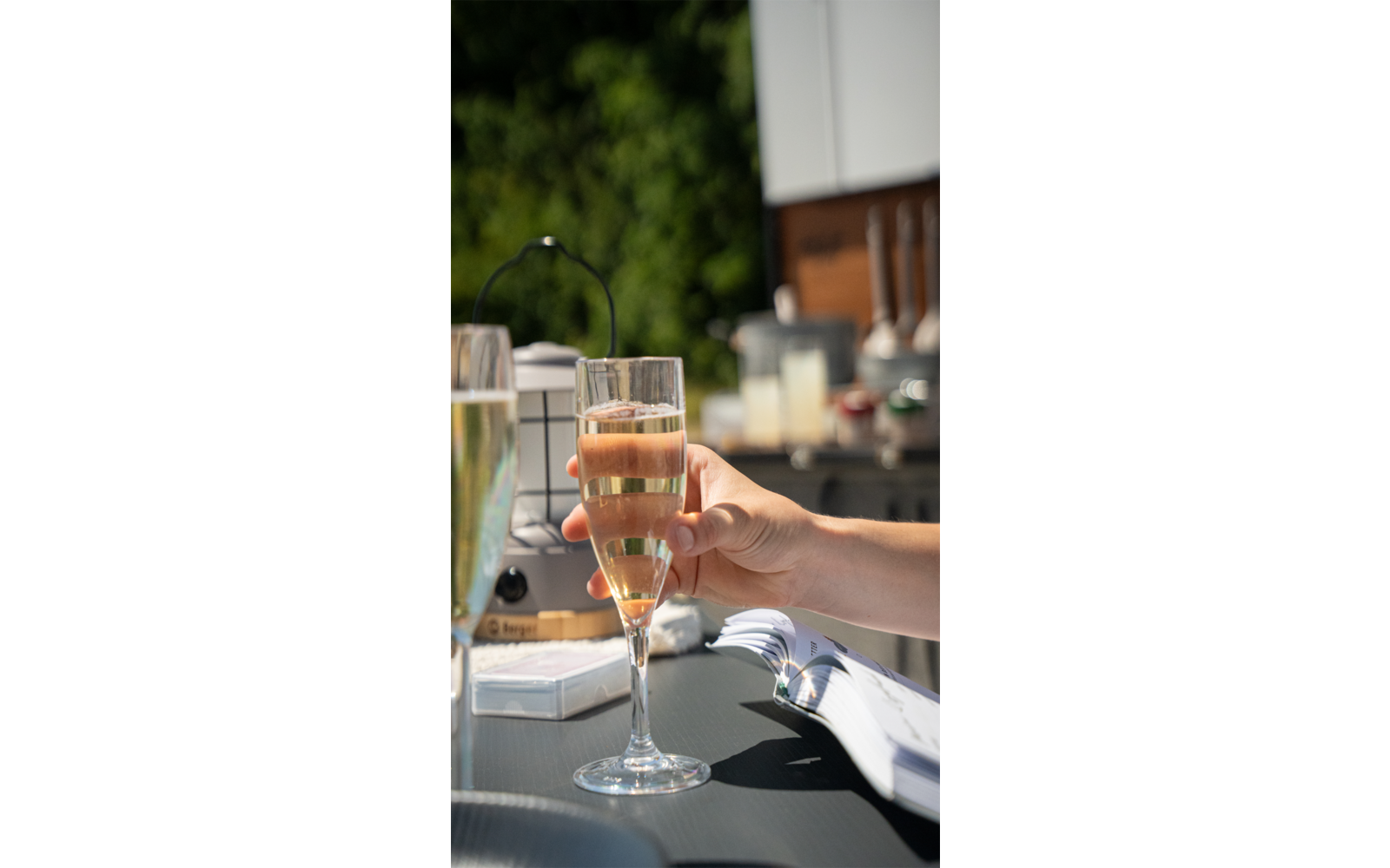 Hand houdt een glas met bruisend drankje vast, op de achtergrond meer glazen en een onscherpe buitenomgeving