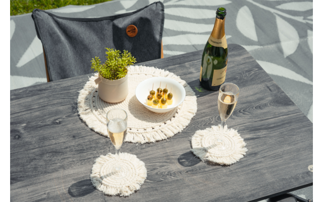 Table en bois gris avec deux flûtes de champagne, une bouteille de champagne ouverte, un bol d'olives sur cure-dents, un petit pot de plante sur un napperon macramé rond, deux sous-verres macramé et une chaise grise en arrière-plan