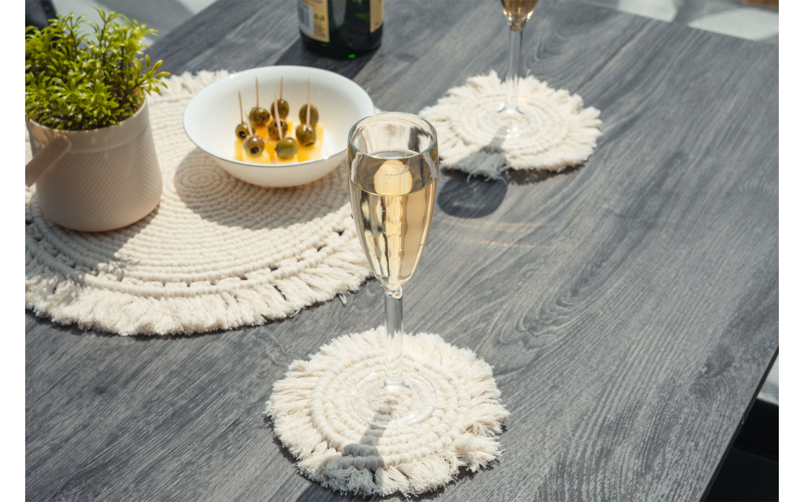 Table en bois grise avec deux dessous de verre macramé, au premier plan une flûte contenant un liquide doré clair, en arrière-plan une autre flûte, un bol d'olives vertes sur des pics et une petite plante en pot
