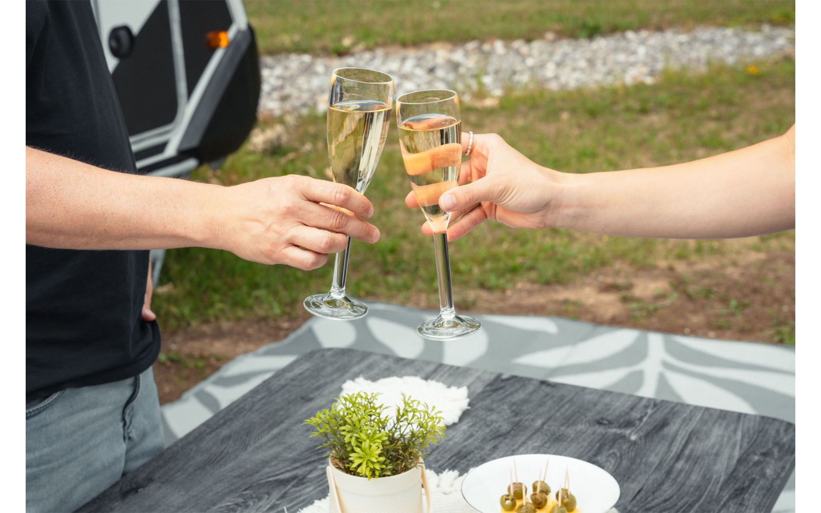 Deux mains trinquant avec des flûtes remplies au-dessus d'une table extérieure ; sur la table un petit pot de plante et une assiette d'olives vertes ; en arrière-plan pelouse, gravier et une caravane