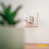 Weiße Steckdose an weißer Wand mit eingestecktem Netzstecker und grünem verschwommenem Pflanzentopf im Vordergrund