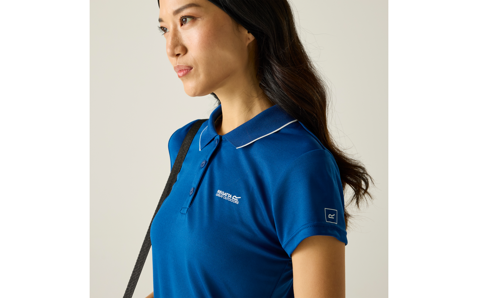 Seitliche Aufnahme einer Person mit dunklen schulterlangen Haaren, blaues Poloshirt mit weißer Paspel am Kragen, kleines weißes Logo auf der Brust, schwarzer Schulterriemen, heller einfarbiger Hintergrund