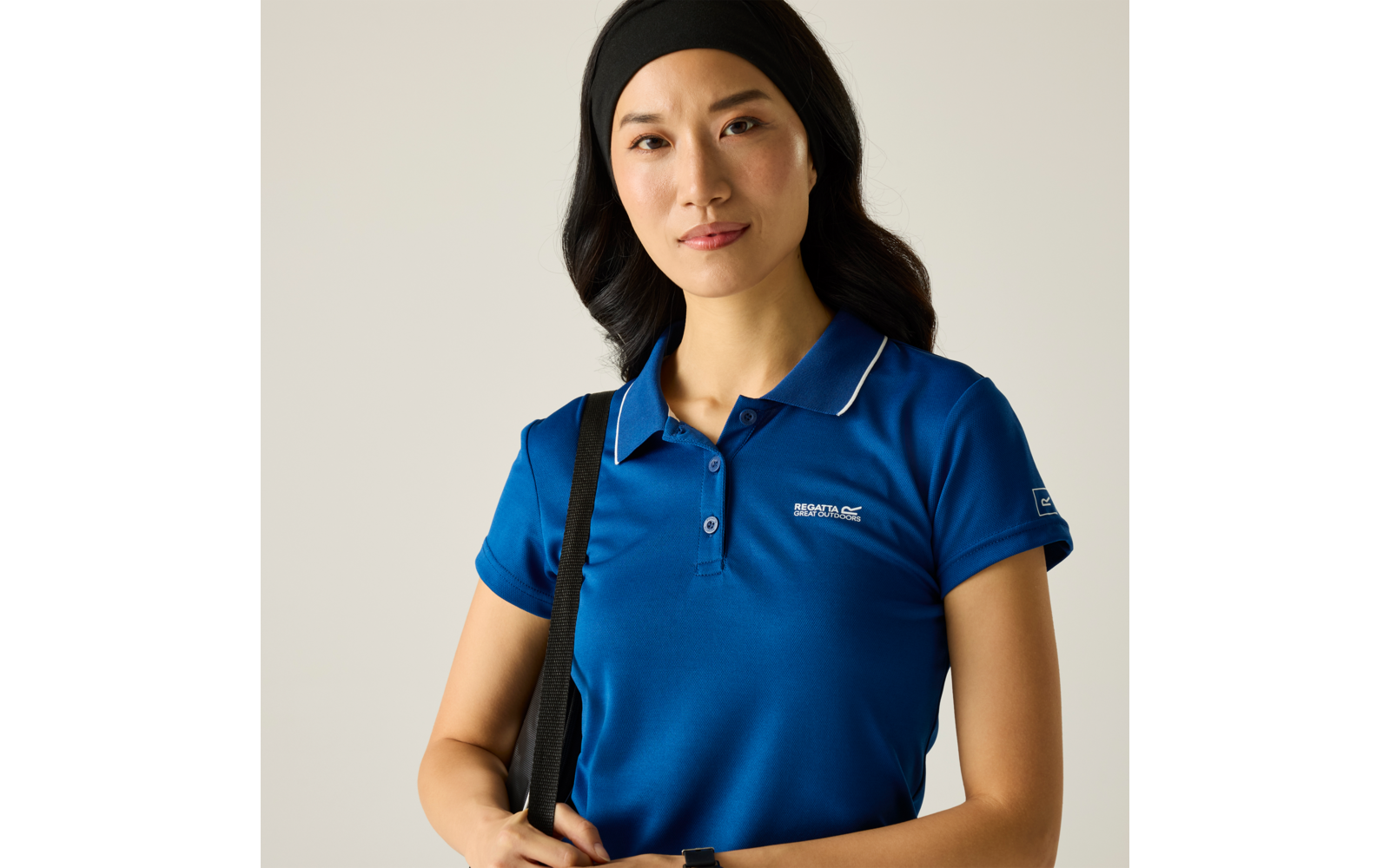 Person mit blauem Poloshirt und schwarzem Stirnband, trägt eine schwarze Umhängetasche, vor hellem einfarbigem Hintergrund