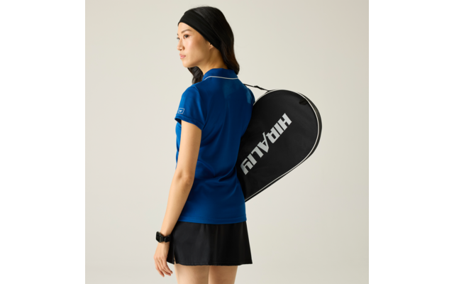 Person in blauem Polohemd, schwarzem Rock und Stirnband trägt eine schwarze Tennistasche mit weißer Schrift über der Schulter, neutraler Hintergrund