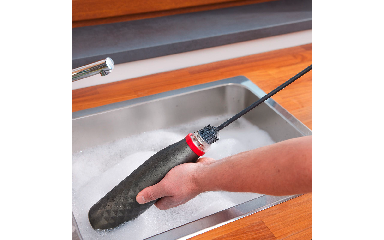 Mano sosteniendo el mango de un cepillo para botellas negro con anillo rojo y cerdas en una varilla larga sobre un fregadero de acero inoxidable lleno de agua jabonosa y espuma