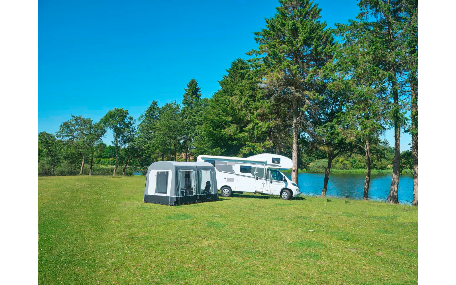 Weißes Wohnmobil neben einem grauen Vorzelt auf grüner Wiese am See, Bäume und blauer Himmel