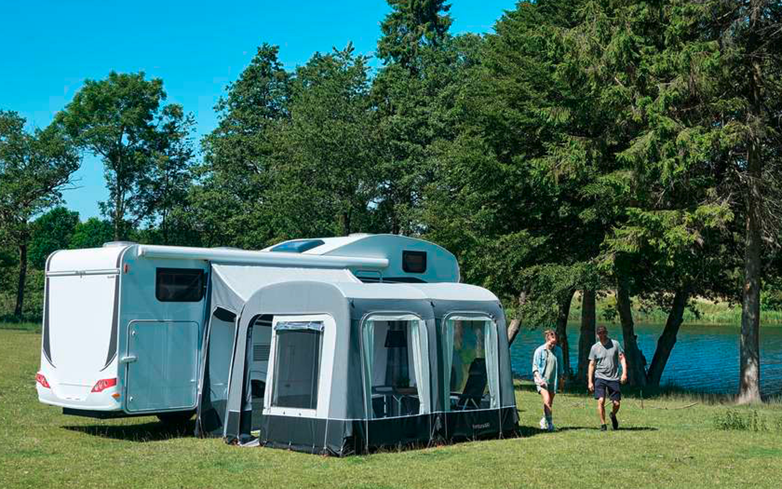 Roulotte bianca con tenda grigia su un prato accanto ad alberi e un lago, due persone camminano sulla riva, cielo azzurro