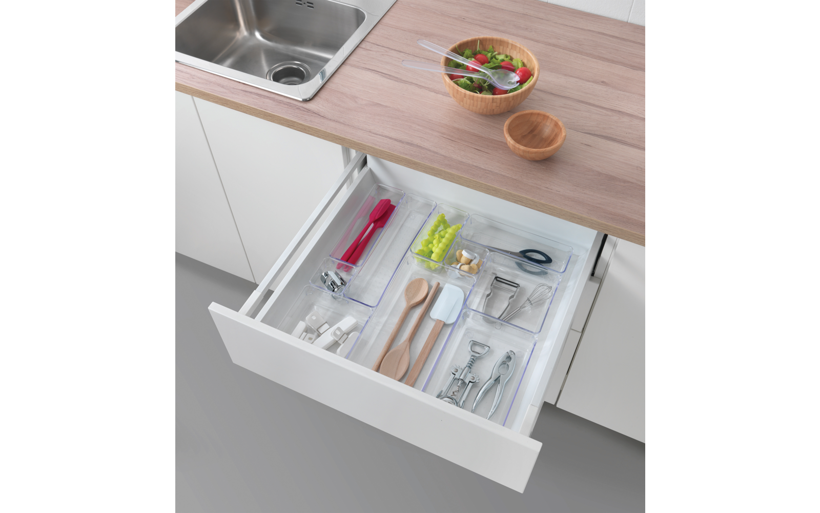 Offene Küchenschublade mit Besteck und Küchenutensilien, darüber Küchenarbeitsfläche mit Spüle und Schalen