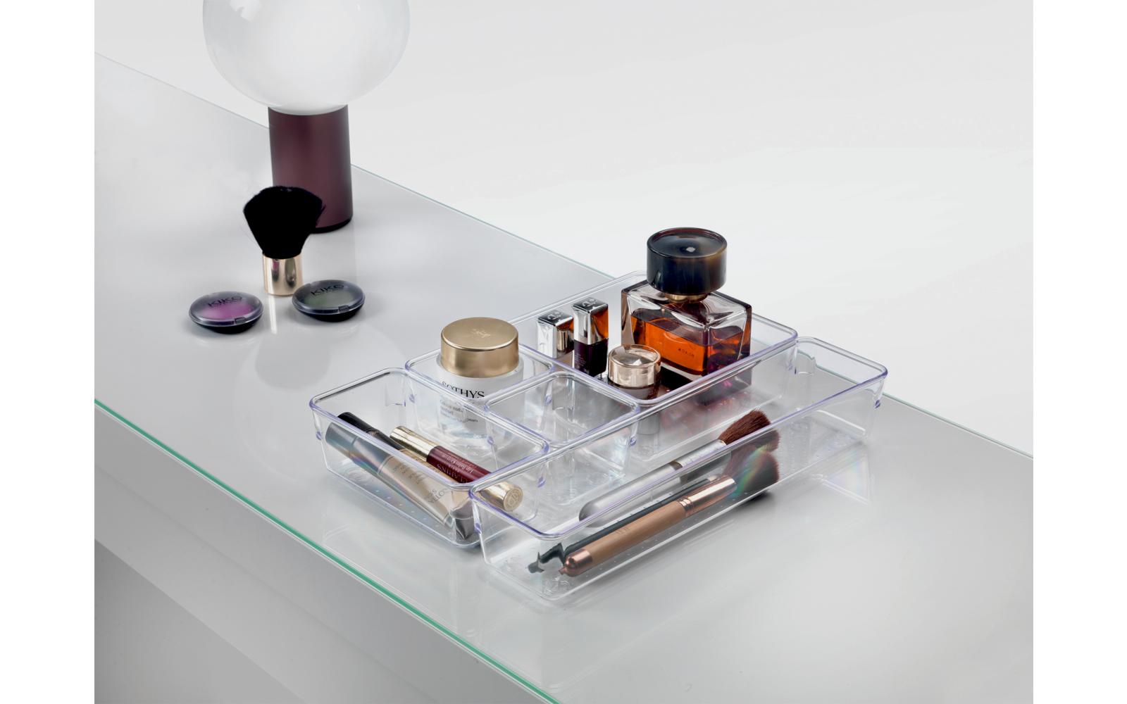 Ein durchsichtiges rechteckiges Tablett mit verschiedenen Kosmetikflaschen und einem runden Make-up-Pinsel auf weißem Untergrund