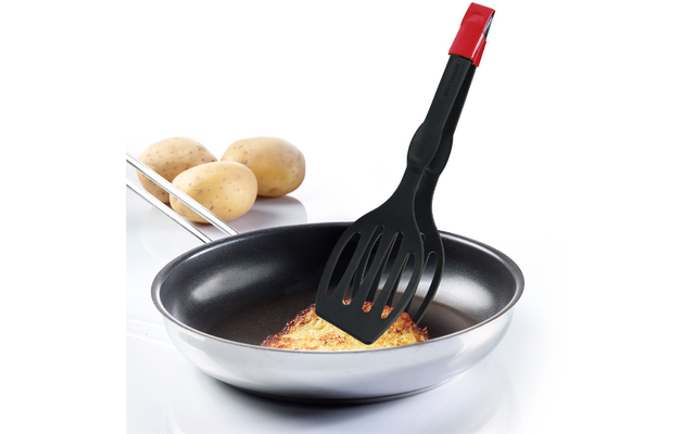 Poêle noire antiadhésive avec un morceau plat doré d'aliment soulevé par une spatule noire fendue au manche rouge ; trois pommes de terre en arrière-plan sur un fond blanc