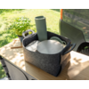 Gray felt basket with handles on a wooden table containing stacked green plant pots, a round silver metal plate, a coiled green hose and white sheets; left a small pot with green leaves; right part of a gray car rear; grass in the background