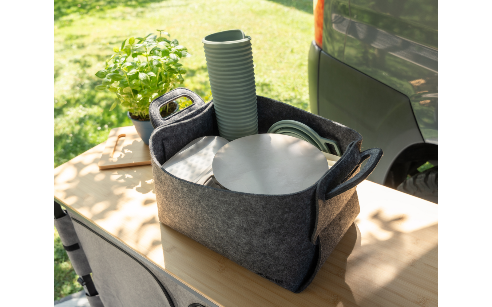 Gray felt basket with handles on a wooden table containing stacked green plant pots, a round silver metal plate, a coiled green hose and white sheets; left a small pot with green leaves; right part of a gray car rear; grass in the background