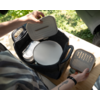 Two hands hold lids above a dark felt basket with handles; inside the basket a round white plate and stacked containers with perforated lids. The basket sits on a wooden table, grass and a car tire visible in the background