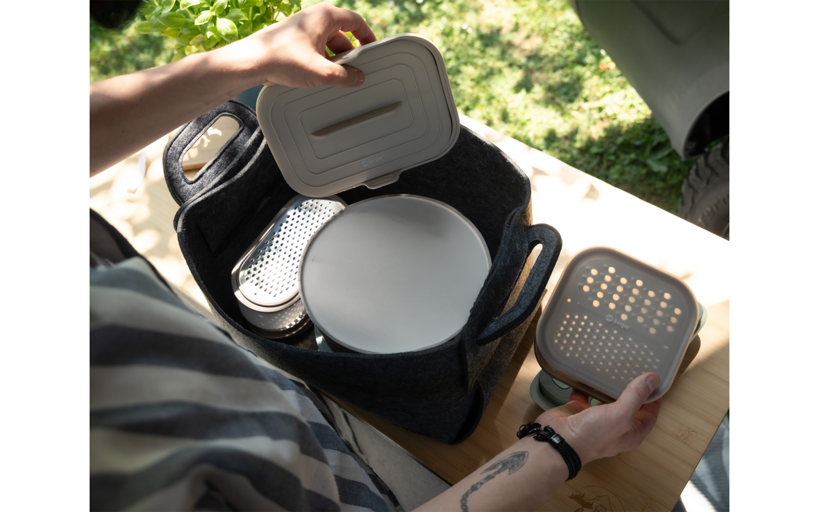 Two hands hold lids above a dark felt basket with handles; inside the basket a round white plate and stacked containers with perforated lids. The basket sits on a wooden table, grass and a car tire visible in the background