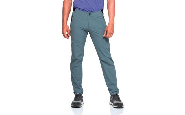 Unterkörper einer stehenden Person von Hüfte bis Fuß, trägt blaugrüne Hose, oberer Saum eines violetten T-Shirts sichtbar, schwarze Sportschuhe mit weißer Sohle, Hände seitlich sichtbar