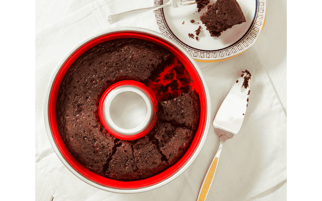 Dark brown round cake in a red ring-shaped pan with one slice removed; the slice on a plate with a fork and crumbs, a cake server beside it on a white tablecloth