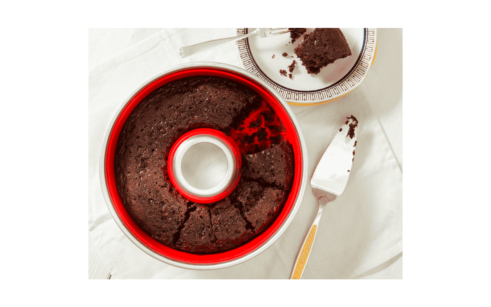Dark brown round cake in a red ring-shaped pan with one slice removed; the slice on a plate with a fork and crumbs, a cake server beside it on a white tablecloth