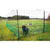 Small black dog inside a low green mesh fence on grass; light-colored dog bed inside