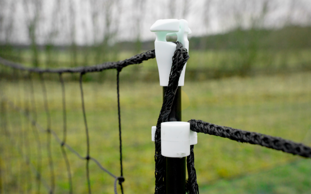 Black braided rope threaded through white plastic clips on a post, fence netting to the left, blurred grassy field in the background