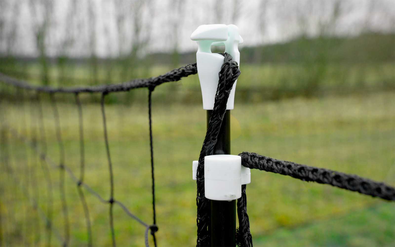 Black braided rope threaded through white plastic clips on a post, fence netting to the left, blurred grassy field in the background