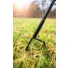 Close-up of a three-pronged hand cultivator with a black handle in yellowish dry grass, blurred trees and sunlight in the background