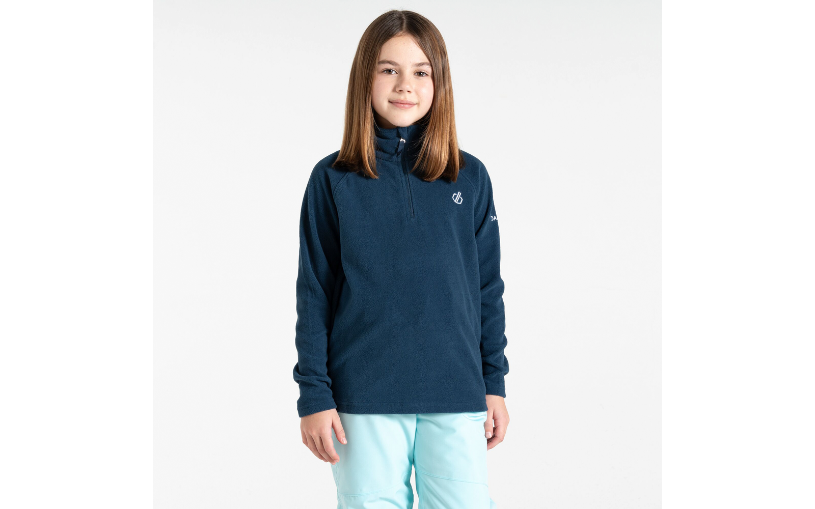 Junges Mädchen steht vor weißem Hintergrund, trägt blauen Pullover und helle Hose