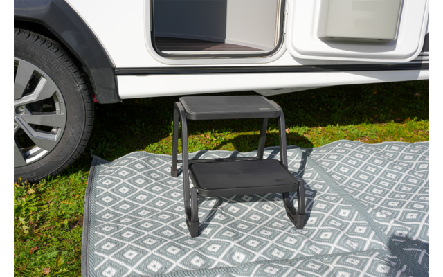 Kleine zweistufige Trittstufe auf gemustertem Außenvorleger vor geöffneter Wohnmobiltür; Fahrzeugrad links sichtbar