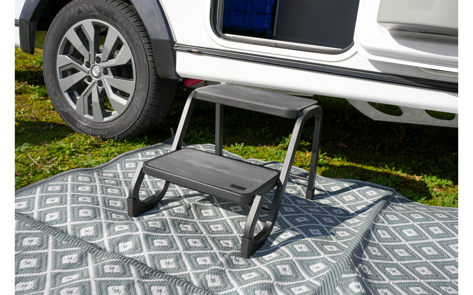 Zweistufige schwarze Trittstufe auf grauem, gemustertem Teppich vor geöffneter Seitentür eines weißen Fahrzeugs; Fahrzeugrad und Gras sichtbar