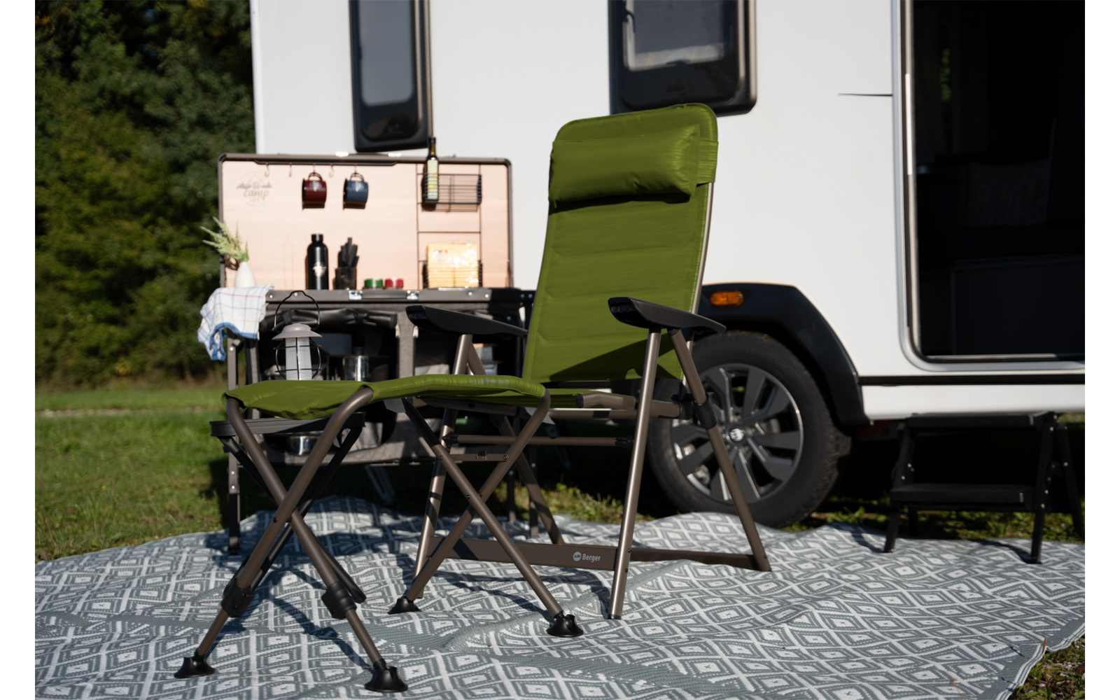 Grüner Klappstuhl mit Klapphocker auf gemusterter Matte vor offenem Wohnmobil; Kasten mit aufgehängten Utensilien, Bäume im Hintergrund