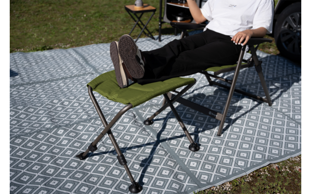 Person sitzt auf grünem Klappstuhl, Füße auf grünem Fußhocker, beide auf grauem, weißgemustertem Outdoor-Teppich; im Hintergrund kleiner Klapptisch, Gras und ein Auto