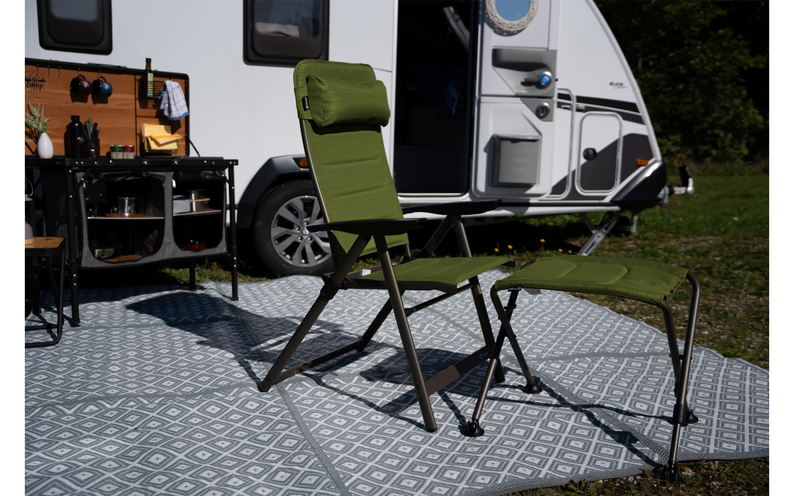 Grüner Campingstuhl mit passendem Fußhocker auf gemusterter Outdoor-Matte vor geöffneter Tür eines Wohnmobils; Outdoor-Küchenzeile links sichtbar