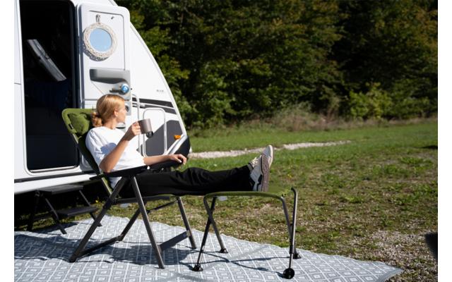 Person mit blondem Haar sitzt auf einem Campingstuhl vor einem Wohnmobil, hält eine Tasse, hat die Füße auf einer Fußablage; Teppich auf Gras, Bäume im Hintergrund