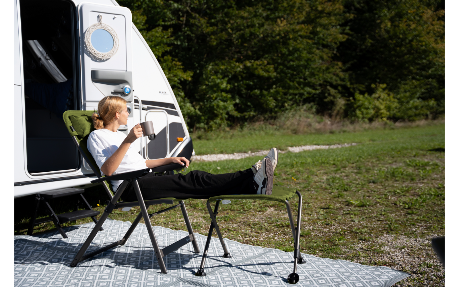 Person mit blondem Haar sitzt auf einem Campingstuhl vor einem Wohnmobil, hält eine Tasse, hat die Füße auf einer Fußablage; Teppich auf Gras, Bäume im Hintergrund