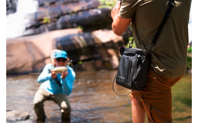Twee personen in ondiep water voor rotsen: één knielt en houdt een vis vast, de ander staat op de voorgrond en maakt een foto; kleine waterval op de achtergrond