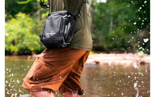 Onderlichaam van een persoon wadend in water, oranje shorts, olijfgroen shirt, zwarte schoudertas, opspattend water, beboste oever op de achtergrond