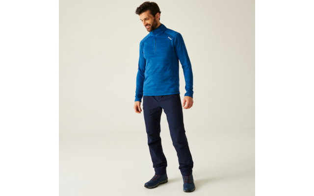 Mann mit blauem Pullover und dunkler Hose steht vor heller Wand