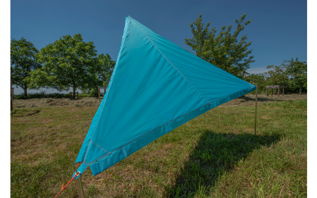 Blaues, dreieckiges Segel vor blauem Himmel auf einer grünen Wiese mit Bäumen im Hintergrund