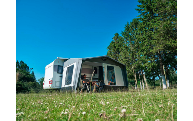 Weißer Wohnwagen mit angebautem Vorzelt auf grüner Wiese; zwei Personen sitzen an einem Tisch im Vorzelt; Bäume rechts; blauer Himmel