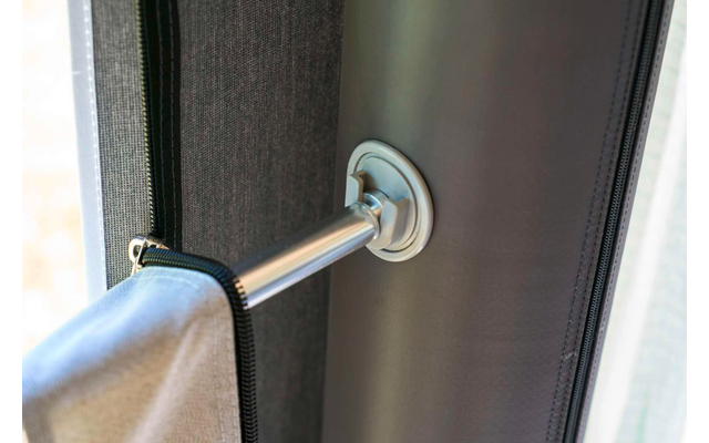 Metallstange mit runder Halterung an einer dunklen vertikalen Fläche; an der Stange hängt ein hellgraues Stoffteil mit sichtbarem Reißverschluss; im Hintergrund heller Fensterbereich