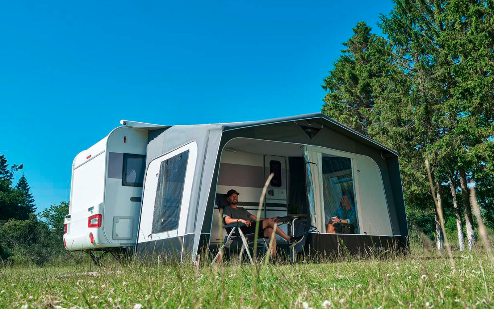 Caravana blanca con toldo anexo en un prado verde; dos personas sentadas en una mesa dentro del toldo; árboles a la derecha; cielo azul