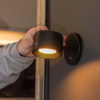 Hand mit Ring und grauem Ärmel hält schwarze zylindrische Wandlampe, die gelbes Licht abstrahlt; Lampenfuß an dunkler Wand montiert